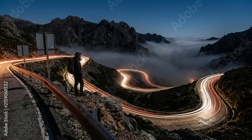 Traveler standing on a scenic winding mountain road at night with long exposure car light trails and dramatic clouds in the valley.