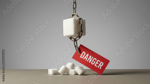 Sugar cube hanging from chain with danger tag above scattered sugar on neutral surface, creating dramatic warning about dietary risk, addiction, and unhealthy consumption