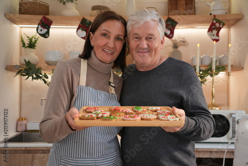 An elderly couple, smiling happily, dressed in aprons, carried wooden trays and baked and decorated gingerbread cookies together to celebrate Christmas with their family.