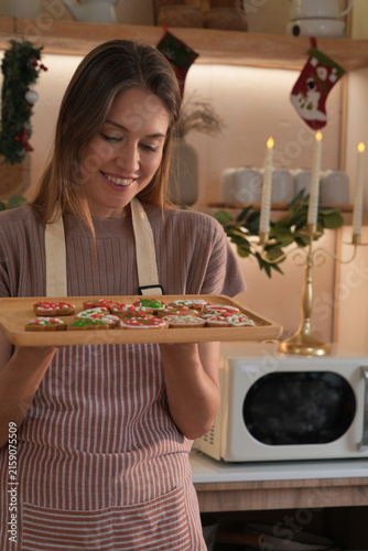 A happy, smiling woman wearing an apron holds a wooden tray filled with beautifully decorated gingerbread cookies, celebrating Christmas.