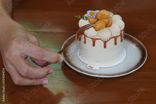 The right hand is holding a fork and cutting a piece of coffee cake and macadamia caramel decorated with purple and yellow flowers, whipped cream, and macadamia nuts on brown and white ceramic dish 