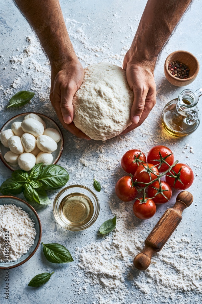 Fototapeta premium Hands arranging pizza ingredients including dough, tomatoes, mozzarella, basil, olive oil and flour on a light kitchen surface, top view flat lay.