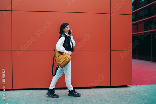 Hispanic woman calling phone mobile roaming connection conversation while walking. Spanish female student traveler with backpack text chatting on cellular near bright red color wall background.