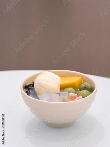 A vertical shot of a ceramic bowl filled with an Asian shaved ice dessert, featuring a scoop of vanilla ice cream, yellow pudding, green mochi balls, tapioca pearls, and jelly cubes on a white table.