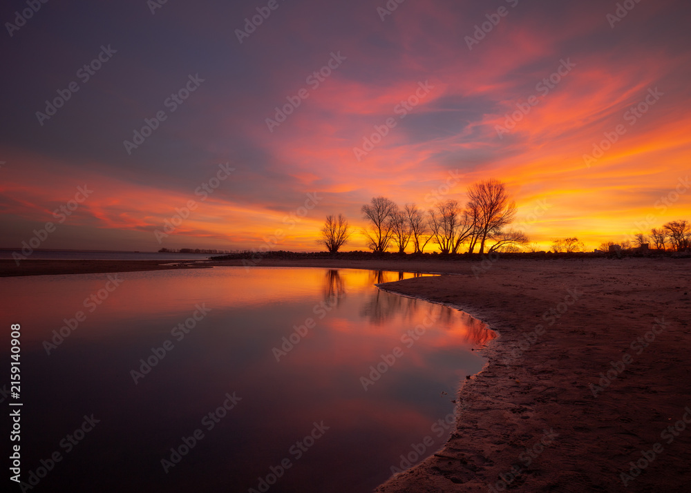 Obraz premium Vivid orange sunset reflected in calm water with bare trees on the shore, Ukraine