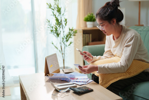 Принти на полотні Woman using smartphone and payment card while reviewing household bills at home,