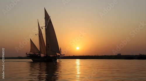 Traditional Sailboat Silhouette Navigating Near Shore with Bright Sun Setting Over Calm Sea
