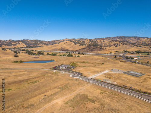 Aerial view showing dry golden fields, rolling hills, a small blue pond, buildings, a highway, and new road construction in Lagoon Valley Park, Vacaville, California