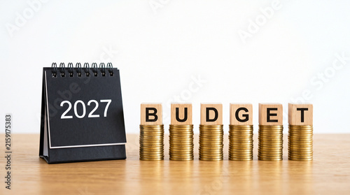 Financial budget planning for 2027, wooden blocks spelling budget on stacks of gold coins, black desk calendar with 2027 year isolated on White Background