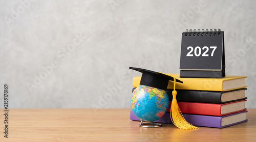 Class of 2027 graduation concept, academic books with mini globe and mortarboard hat, 2027 calendar on wooden table isolated on White Background