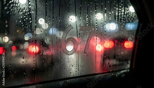 Rain-covered car window with the number 67 visible on the glass, blurred city street lights and traffic forming a moody urban background, capturing rainy weather, travel, reflection and cinematic vibe