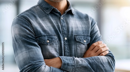 Confident man in a denim shirt, arms crossed, in a modern office environment.