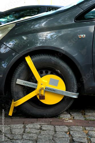 Photography Bright yellow wheel clamp secured around a parked car tire on a paved street, showing vehicle immobilization, parking enforcement, security restriction