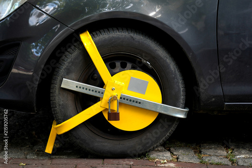 Konstfotografi Bright yellow wheel clamp secured around a parked car tire on a paved street, showing vehicle immobilization, parking enforcement, security restriction