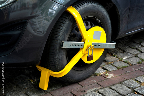 Fotografía Bright yellow wheel clamp secured around a parked car tire on a paved street, showing vehicle immobilization, parking enforcement, security restriction