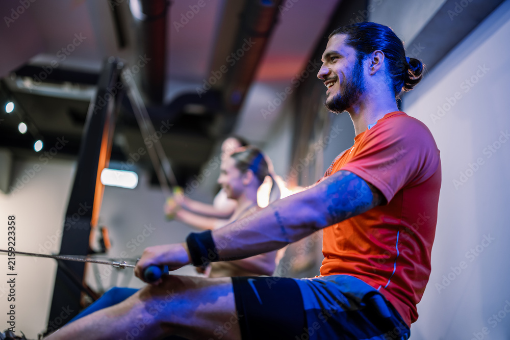 Fototapeta premium Side profile view of a muscular man focused on his form while exercising on a rowing ergometer next to other people in a dark, atmospheric gym.