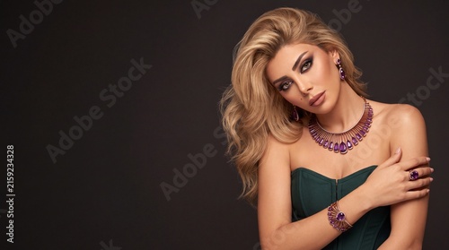 Blonde woman wearing rose gold purple amethyst gemstone sunburst fringe jewelry set in green corset dress, model portrait with copy space