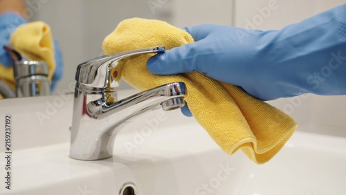 Person in blue gloves cleaning a chrome bathroom faucet with a yellow microfiber cloth.