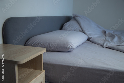 In the left corner of the frame, a part of a wooden pedestal is visible, and on the gray bed there are two pillows and a blanket spread out. The concept of the comfort of a sleeping place, preparation