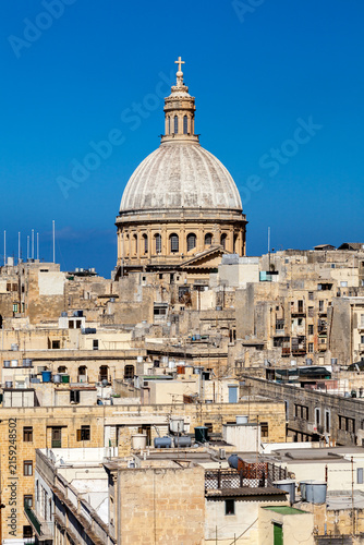 The dome of the Carmelite Basilica rises above the dense rooftops of Valletta, Malta, against a clear blue sky