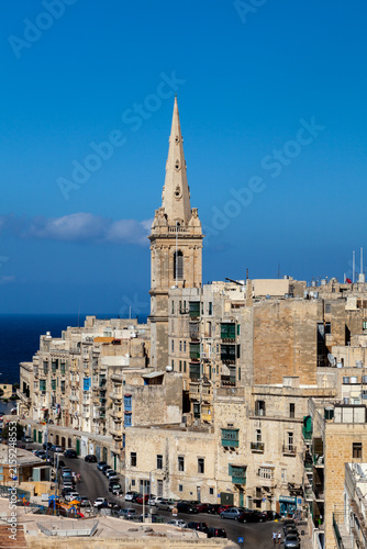St Paul's Pro-Cathedral spire rises above Valletta's rooftops and traffic-lined streets, Malta, with the sea visible beyond