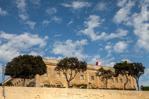 A long limestone building with a Maltese flag sits atop a fortified wall beneath a dramatic cloud-filled sky in Valletta