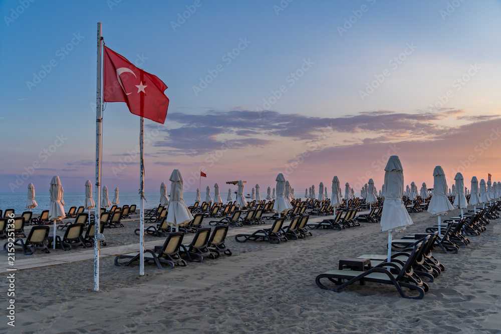Fototapeta premium Beach chairs and umbrellas beneath a Turkish flag during a Mediterranean sunset in Antalya, Turkey.