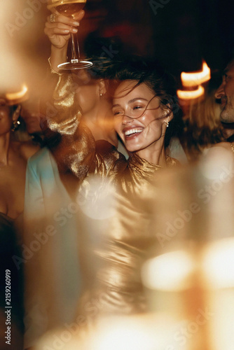 Smiling woman in a shimmering gold dress raises a wine glass amid a glamorous candlelit nighttime celebration. Motion blur, selective focus. 