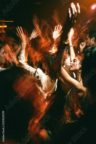 Crowded nightclub dancers raise their hands under warm orange lights, captured with energetic motion blur from a close perspective. Motion blur, selective focus.