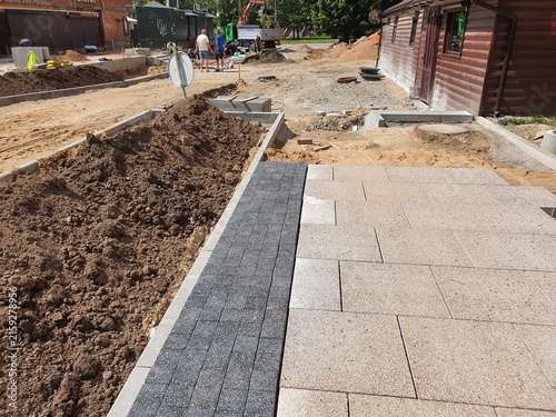 An outdoor construction site showing sidewalk and pavement installation work in progress. In the foreground, newly laid light gray concrete paving stones are visible, bordered by a strip of darker