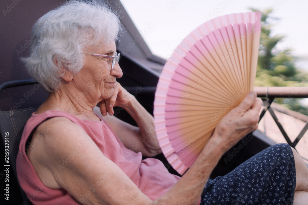 Fototapeta premium Elderly woman cooling herself with a fan while sitting on the balcony during a heatwave