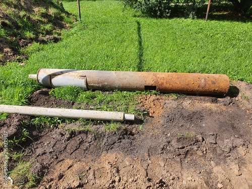 Outdoor construction or utility work scene showing excavated earth with exposed pipes. A large rusty metal pipe with a rectangular cutout lies on disturbed soil next to a white PVC pipe. The ground