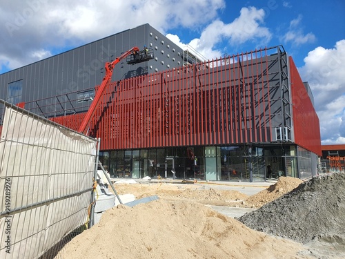 Modern commercial building under construction with distinctive red vertical slat facade and dark gray paneling. An orange articulated boom lift with workers in a basket is positioned against the