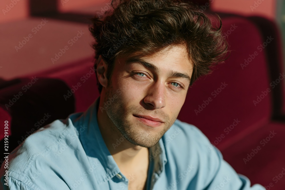 Obraz premium Closeup warm sunlight portrait of relaxed young man with blue eyes on velvet sofa for casual lifestyle headshot background
