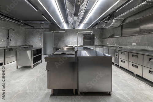 A modern, professional commercial kitchen layout featuring an island cooking suite and extensive stainless steel prep and storage counters against grey marble-tiled walls