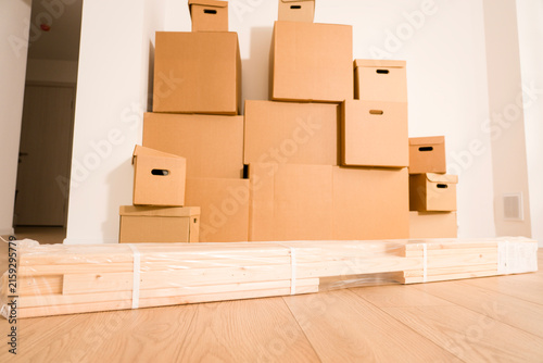 Stacked cardboard boxes fill a room. Various-sized boxes create a storage display. Wooden pallet rests on light wood flooring. White walls and a hallway entrance complete the setting