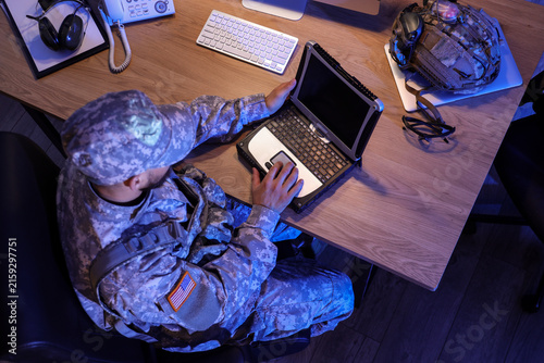 Soldier working with military laptop on desk in office at night, top view