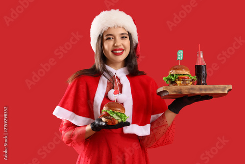 Pretty young woman in Santa costume with tasty burgers and cola on red backgr...