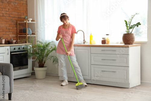 Pretty young woman with headphones and broom cleaning floor in kitchen