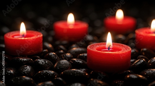 Four red tealight candles burn on dark wet pebbles. Small flames glow in dim light. Warm fire flickers on black stones. Calm atmosphere for spa wellness romantic dinner. Peaceful zen mood with red