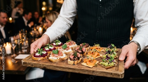 Professional waiter holding a wooden board with delicious gourmet appetizers and salmon canapes at a high end restaurant dinner party or luxury catering event representing hospitality and fine foods.