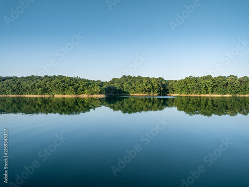 Принти на полотні A still lake mirrors a dense green forest under a cloudless clear blue sky, forming a symmetrical natural landscape