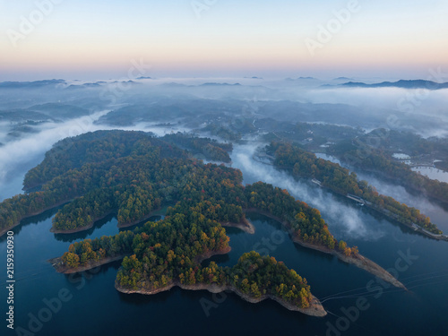 Fotografie Aerial shot of forested islands in a misty lake at early dawn, with distant mountain ranges in the haze
