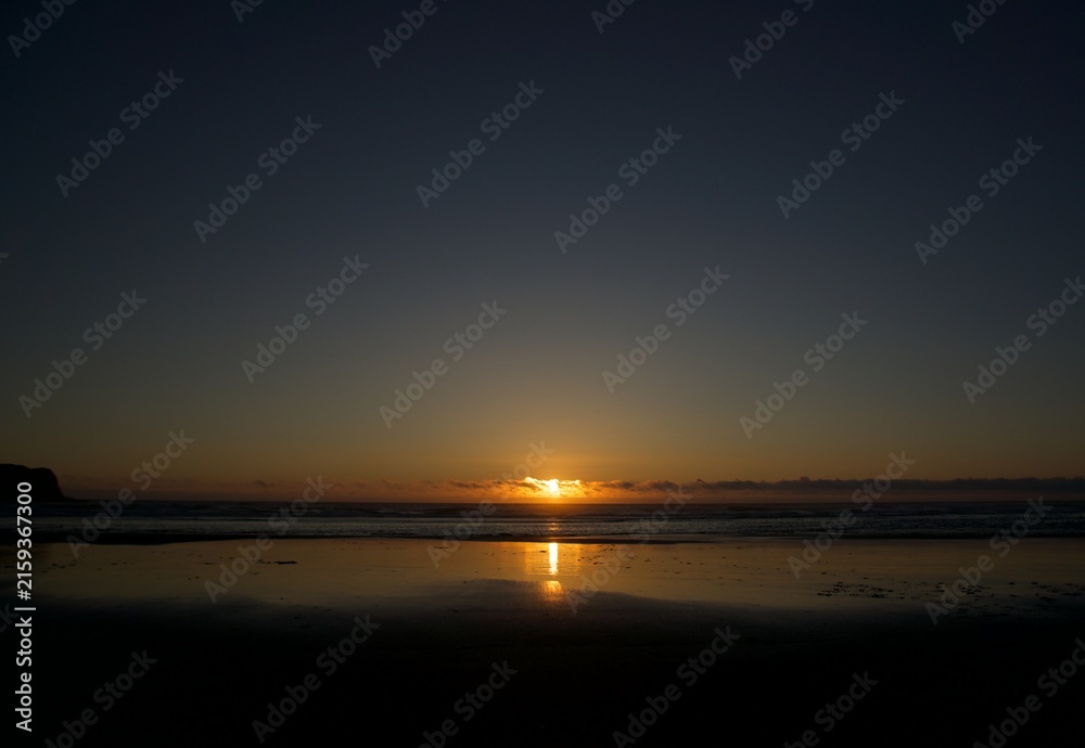 Fototapeta premium Wide Angle Landscape of Oregon Coast at Sunset with Sun Dipping Below Low Cloud Horizon and Perfect Light Beam Reflection on Wet Flat Sandy Beach