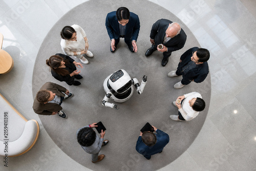 Top view of a small robot surrounded by a diverse team of professionals in business attire, gathered around a circular rug in a bright modern atrium with glass floors for a collaborative discussion.