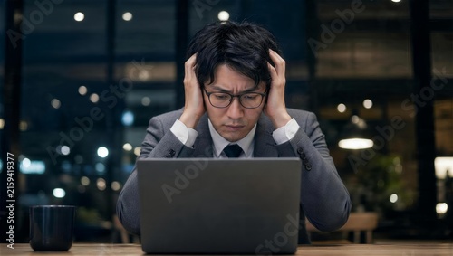 暗い窓辺でノートパソコンを前に頭を抱える眼鏡の会社員男性