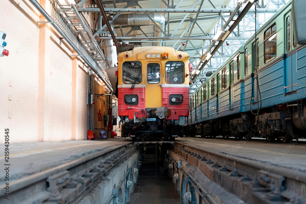 Obraz premium A vibrant yellow and red train sits on the tracks in a busy transit depot, with another blue train awaiting its turn in the background under bright lights