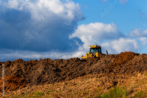 Yellow bulldozer operating on a mound of raw earth at a construction site against a dramatic blue cloudy sky during a sunny day of heavy industrial development work