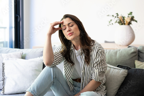 Young woman feeling intense headache or migraine, sitting on a comfortable sofa in a bright modern living room, experiencing stress and exhaustion from the discomfort