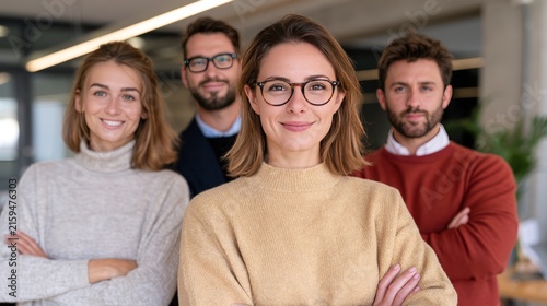 A confident team of four professionals, two women and two men, posing in an office environment. The two women in the foreground are smiling at the camera, one with crossed arms and wearing glasses.